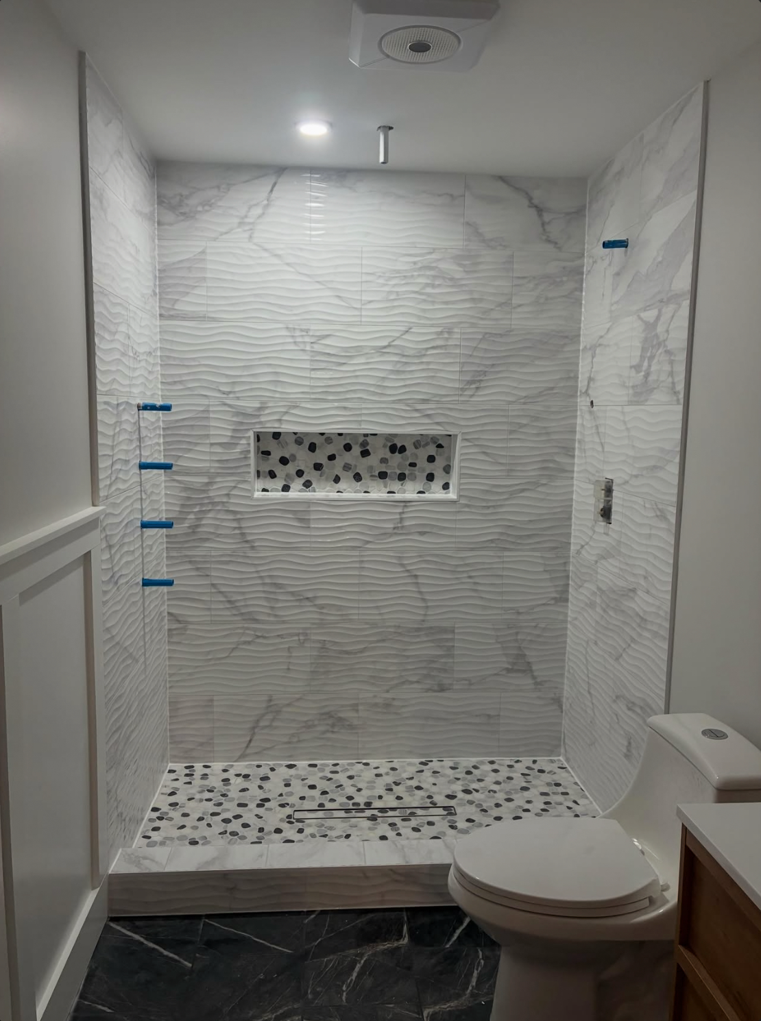 Walk-in shower with wave-textured marble-look tile, pebble mosaic floor, and recessed niche